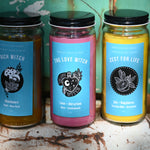 Three of the Spell Candles with Herbs and Crystals all in front of a teal background and on a white rusty surface.