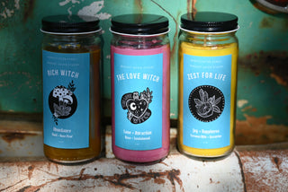 Three of the Spell Candles with Herbs and Crystals all in front of a teal background and on a white rusty surface.