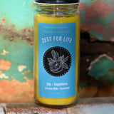 The Zest for Life Spell Candle with Herbs and Crystals in front of a teal background and on a white rusty surface.