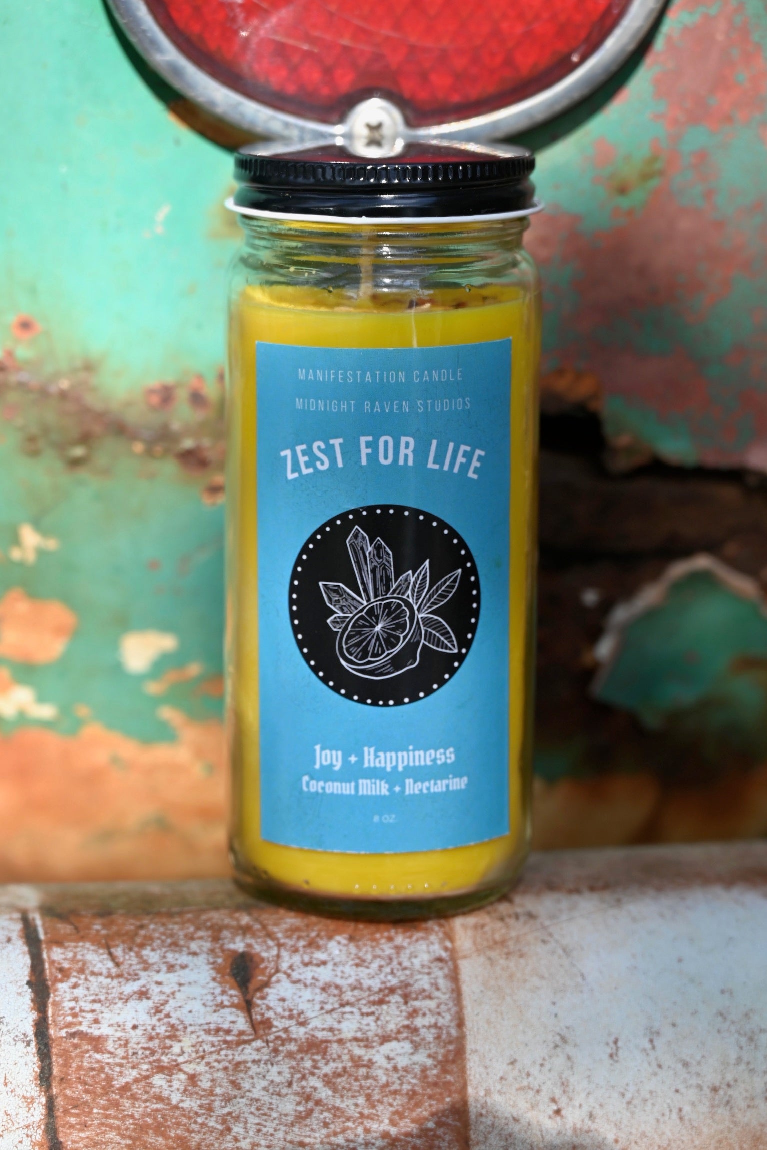 The Zest for Life Spell Candle with Herbs and Crystals in front of a teal background and on a white rusty surface.