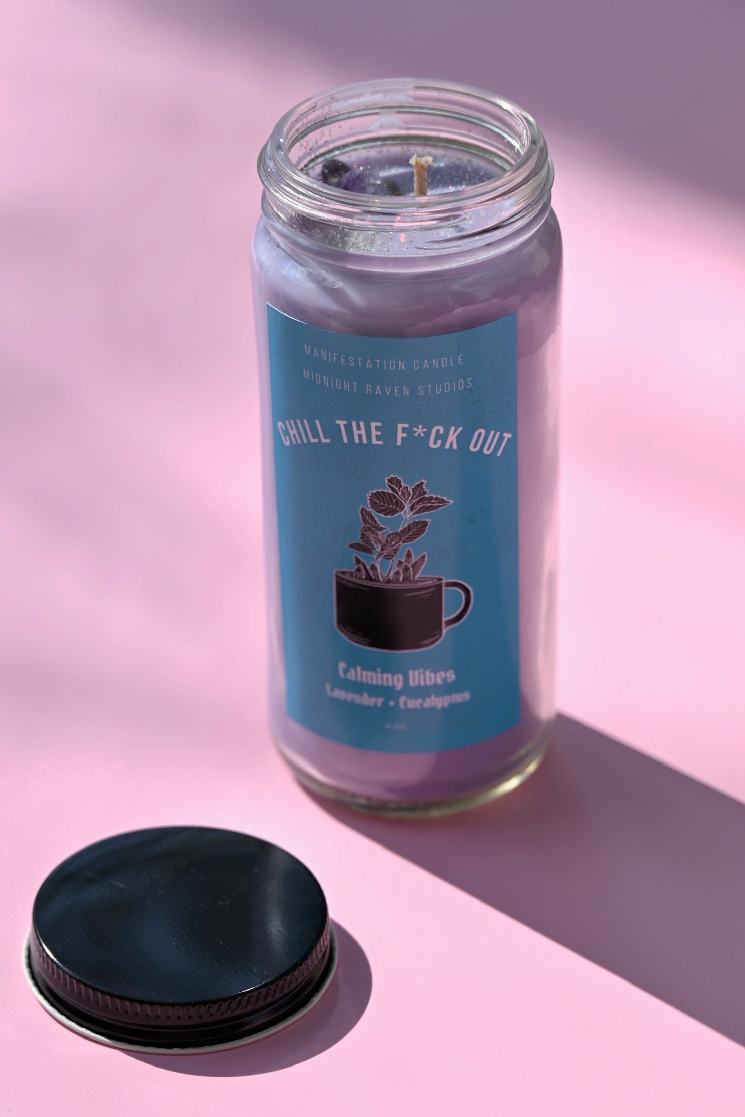 The Chill the F*ck Out Spell Candle with Herbs and Crystals on a pink background with its lid off and to the side.