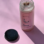 The Goddess Manifestation Candle with Herbs and Crystals on a pink background with its lid to the side.