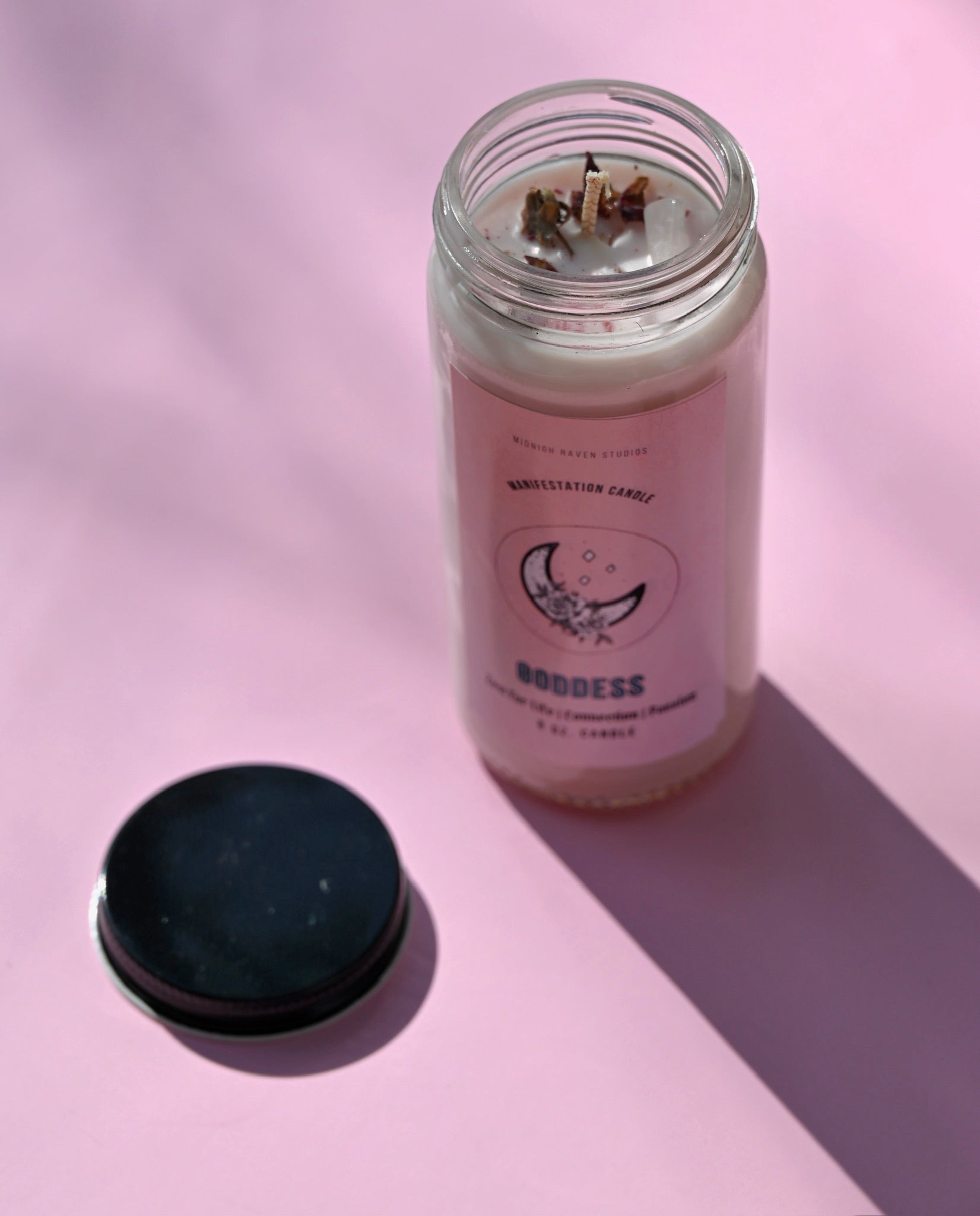 The Goddess Manifestation Candle with Herbs and Crystals on a pink background with its lid to the side.