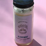 The Alchemist Manifestation Candle with Herbs and Crystals on a pink background.
