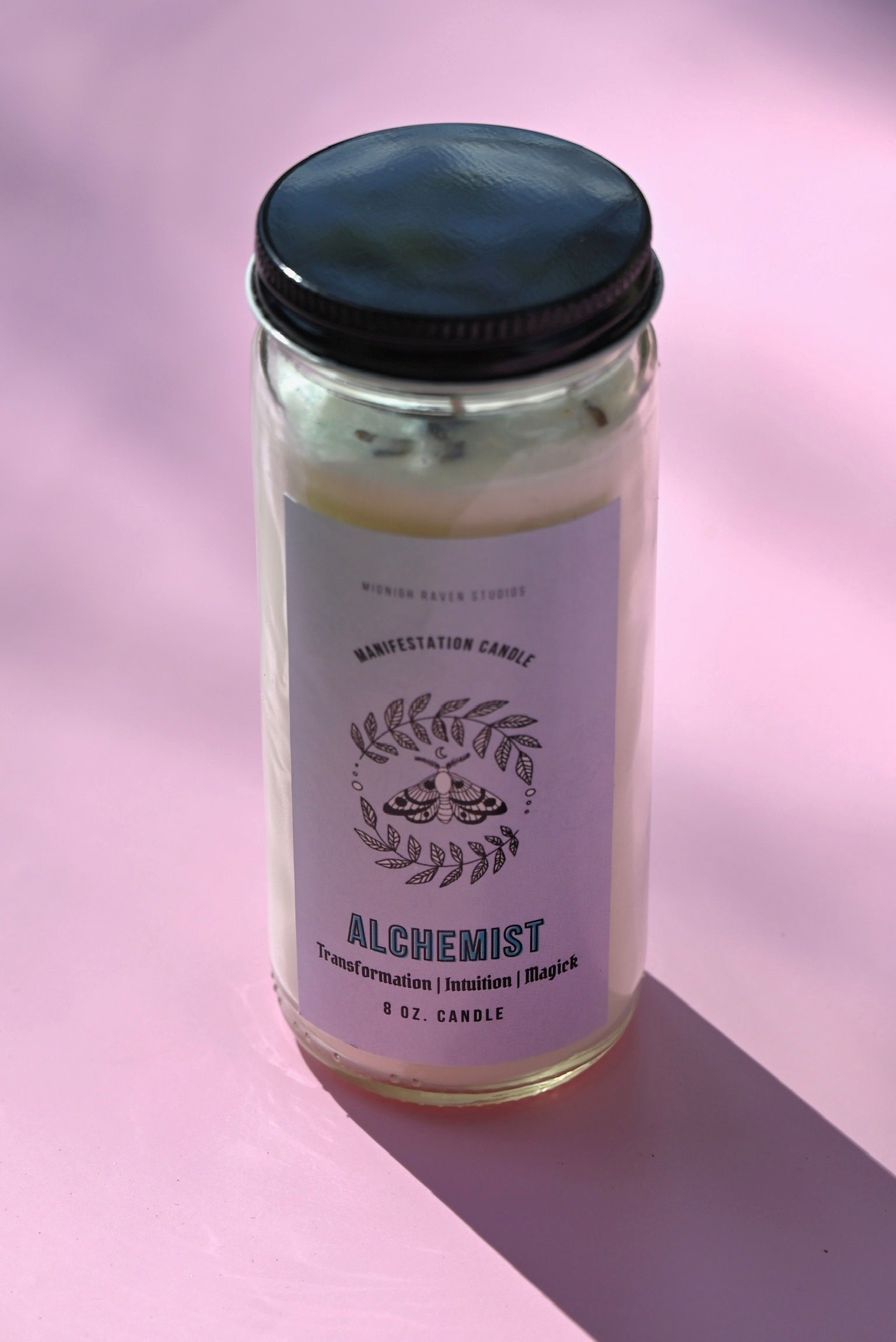The Alchemist Manifestation Candle with Herbs and Crystals on a pink background.