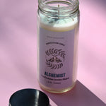 The Alchemist Manifestation Candle with Herbs and Crystals on a pink background with its lid off and to the side.