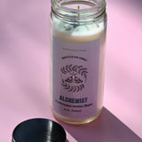 The Alchemist Manifestation Candle with Herbs and Crystals on a pink background with its lid off and to the side.