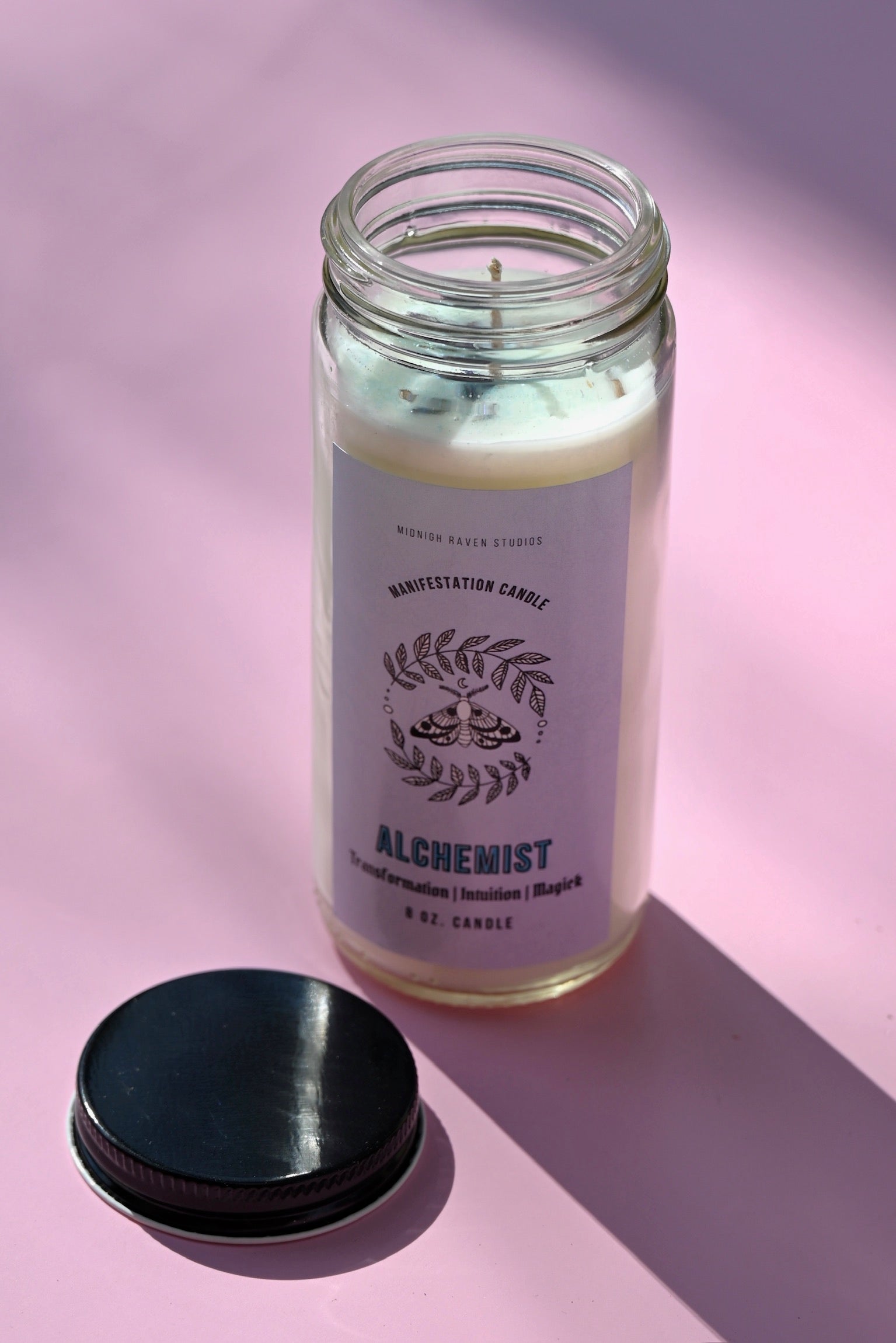 The Alchemist Manifestation Candle with Herbs and Crystals on a pink background with its lid off and to the side.
