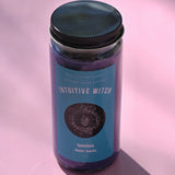 The Intuitive Witch Spell Candle with Herbs and Crystals on a pink background.