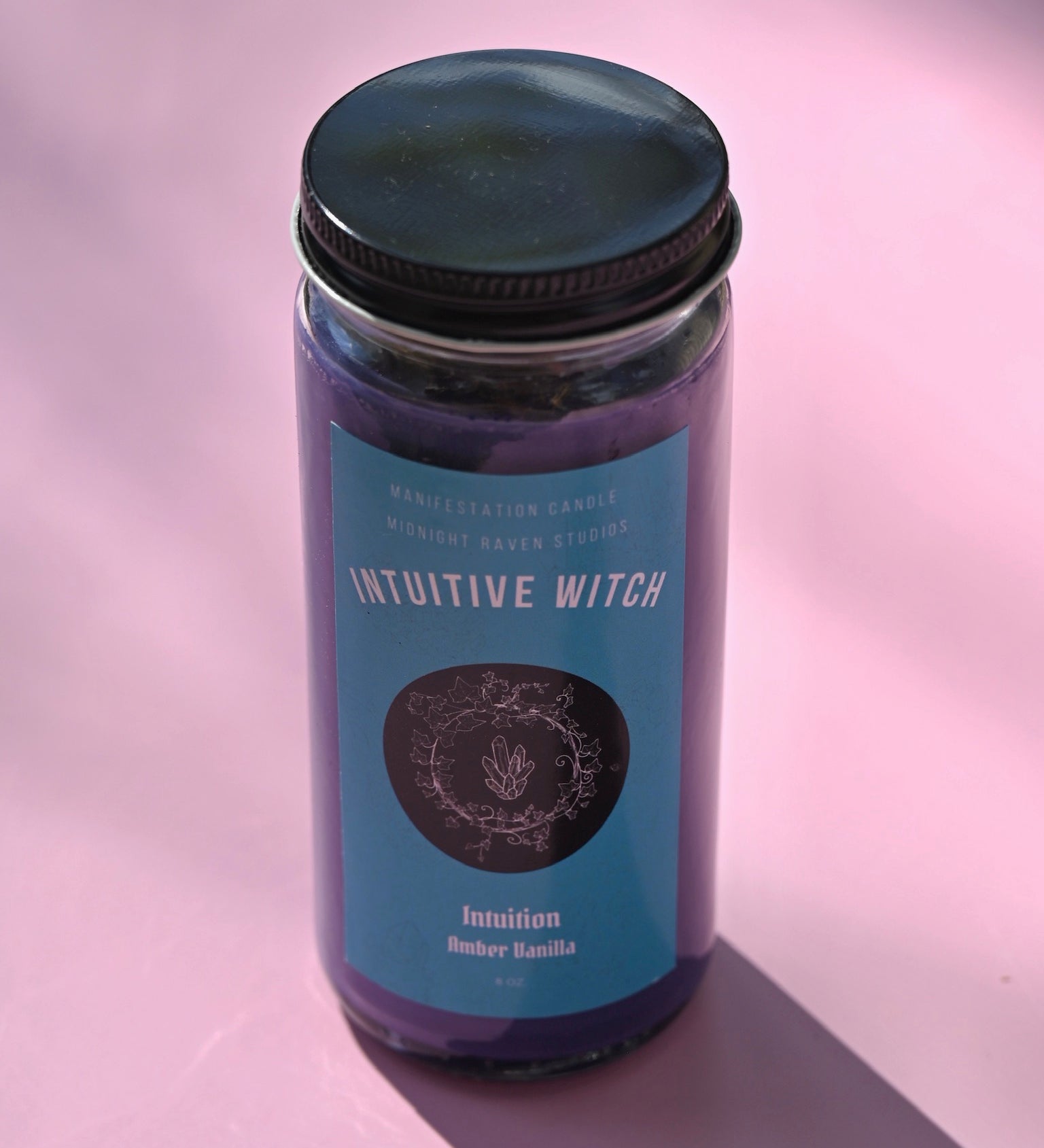 The Intuitive Witch Spell Candle with Herbs and Crystals on a pink background.