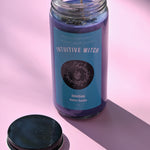 The Intuitive Witch Spell Candle with Herbs and Crystals on a pink background with the lid off and to the side.