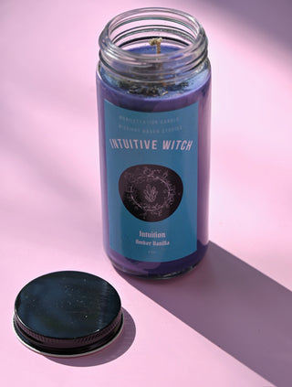 The Intuitive Witch Spell Candle with Herbs and Crystals on a pink background with the lid off and to the side.