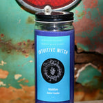 The Intuitive Witch Spell Candle with Herbs and Crystals in front of a teal background and on a white rusty surface.