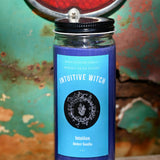 The Intuitive Witch Spell Candle with Herbs and Crystals in front of a teal background and on a white rusty surface.
