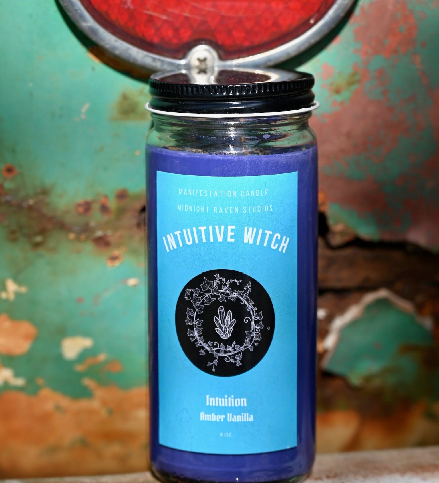 The Intuitive Witch Spell Candle with Herbs and Crystals in front of a teal background and on a white rusty surface.