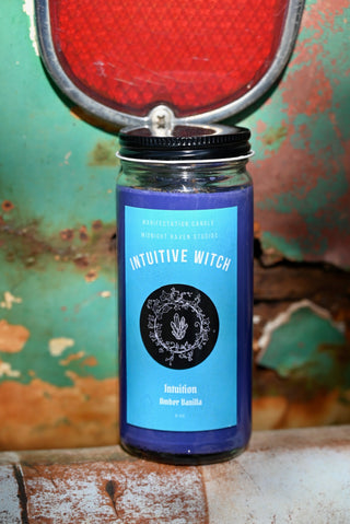 The Intuitive Witch Spell Candle with Herbs and Crystals in front of a teal background and on a white rusty surface.