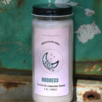 The Goddess Manifestation Candle with Herbs and Crystals in front of a teal background and on a white rusty surface.