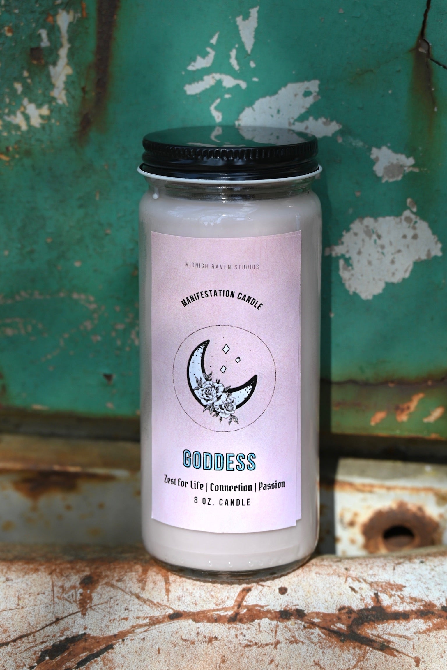 The Goddess Manifestation Candle with Herbs and Crystals in front of a teal background and on a white rusty surface.