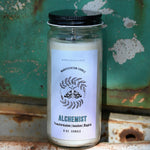 The Alchemist Manifestation Candle with Herbs and Crystals in front of a teal background and on a white rusty surface.
