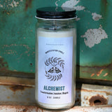 The Alchemist Manifestation Candle with Herbs and Crystals in front of a teal background and on a white rusty surface.