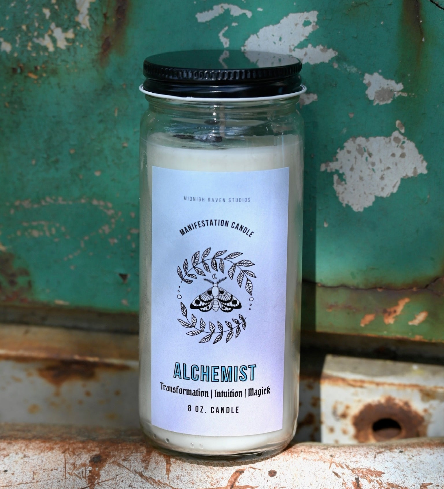 The Alchemist Manifestation Candle with Herbs and Crystals in front of a teal background and on a white rusty surface.