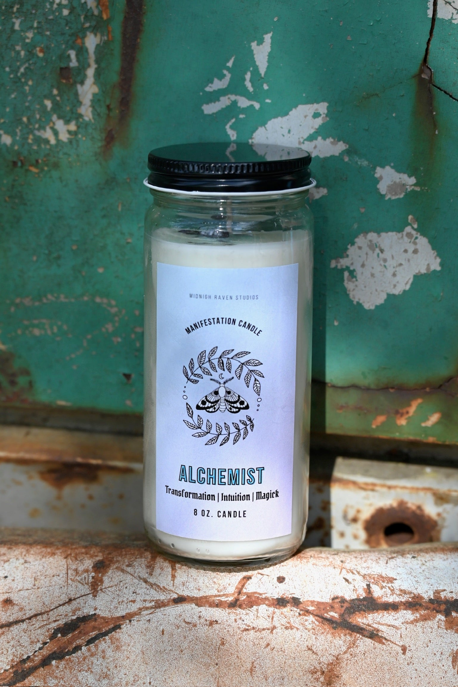 The Alchemist Manifestation Candle with Herbs and Crystals in front of a teal background and on a white rusty surface.