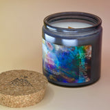 The Cosmic Creator Intention Candle on a a pink and yellow background with the cork lid off and lying flat next to the jar.