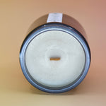 The Moons Ritual Candle on a pink and yellow background lying on its side showing off the wooden wick and gray sparkles in the candle.