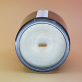 The Moons Ritual Candle on a pink and yellow background lying on its side showing off the wooden wick and gray sparkles in the candle.