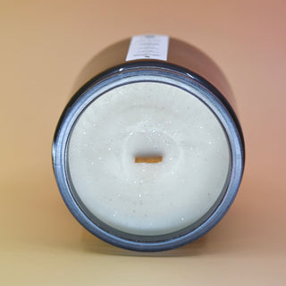 The Moons Ritual Candle on a pink and yellow background lying on its side showing off the wooden wick and gray sparkles in the candle.