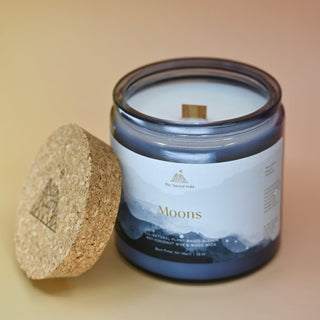 The Moons Ritual Candle on a pink and yellow background with the cork lid off and leaning against the candle jar.