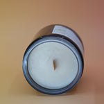 The Moons Ritual Candle on a pink and yellow background lying on its side showing off the wooden wick and gray sparkles in the candle.
