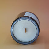The Moons Ritual Candle on a pink and yellow background lying on its side showing off the wooden wick and gray sparkles in the candle.