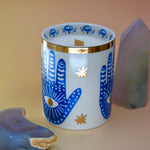 The Hamsa Candle on a pink and yellow background with an agate tower on the right of the candle and an agate crystal moon on the left.