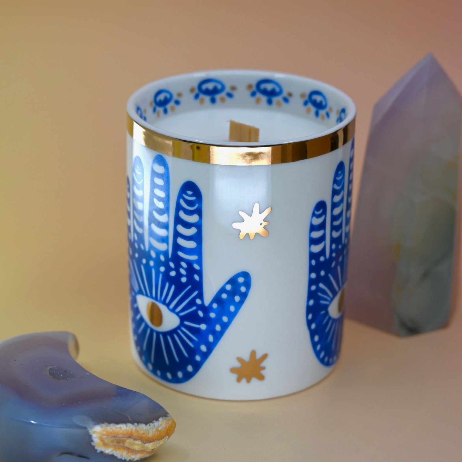 The Hamsa Candle on a pink and yellow background with an agate tower on the right of the candle and an agate crystal moon on the left.