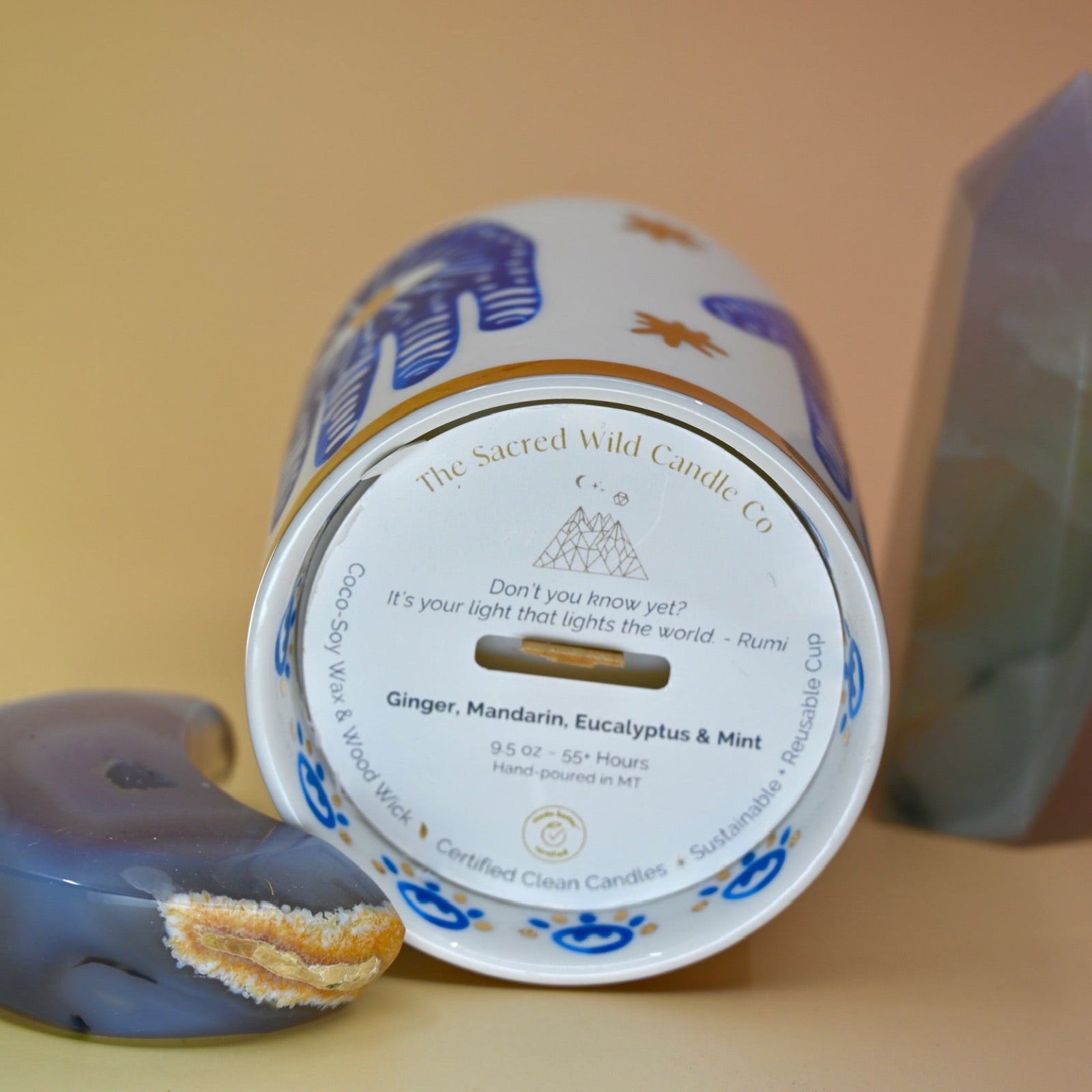 The Hamsa Candle lying down on a pink and yellow background with an agate tower on the right of the candle and an agate crystal moon on the left.