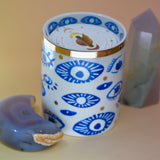 The "Evil Eye" Candle on a pink and yellow background with an agate tower on the right of the candle and an agate crystal moon on the left.