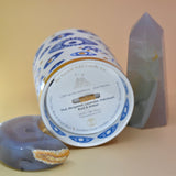 The "Evil Eye" Candle lying down on a pink and yellow background with an agate tower on the right of the candle and an agate crystal moon on the left.