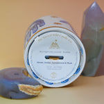 The Hamsa Candle lying down on a pink and yellow background with an agate tower on the right of the candle and an agate crystal moon on the left.