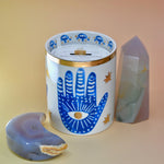 The Hamsa Candle on a pink and yellow background with an agate tower on the right of the candle and an agate crystal moon on the left.