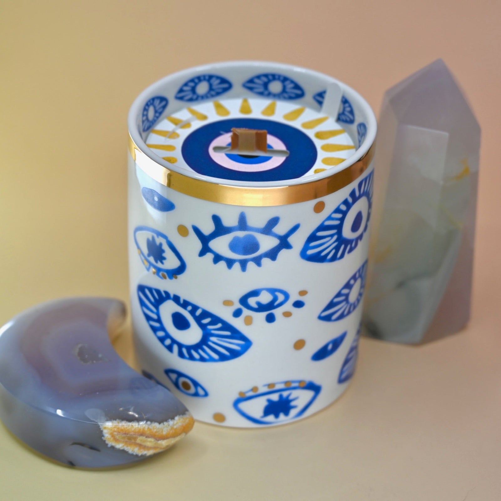 The "Evil Eye" Candle on a pink and yellow background with an agate tower on the right of the candle and an agate crystal moon on the left.