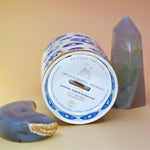 The "Evil Eye" Candle lying down on a pink and yellow background with an agate tower on the right of the candle and an agate crystal moon on the left.