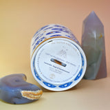 The "Evil Eye" Candle lying down on a pink and yellow background with an agate tower on the right of the candle and an agate crystal moon on the left.