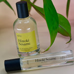The Hinoki Sesame 5ml Unisex Eau de Parfum regular sized bottle and the Travel Sprayer on a pink background with leaves.