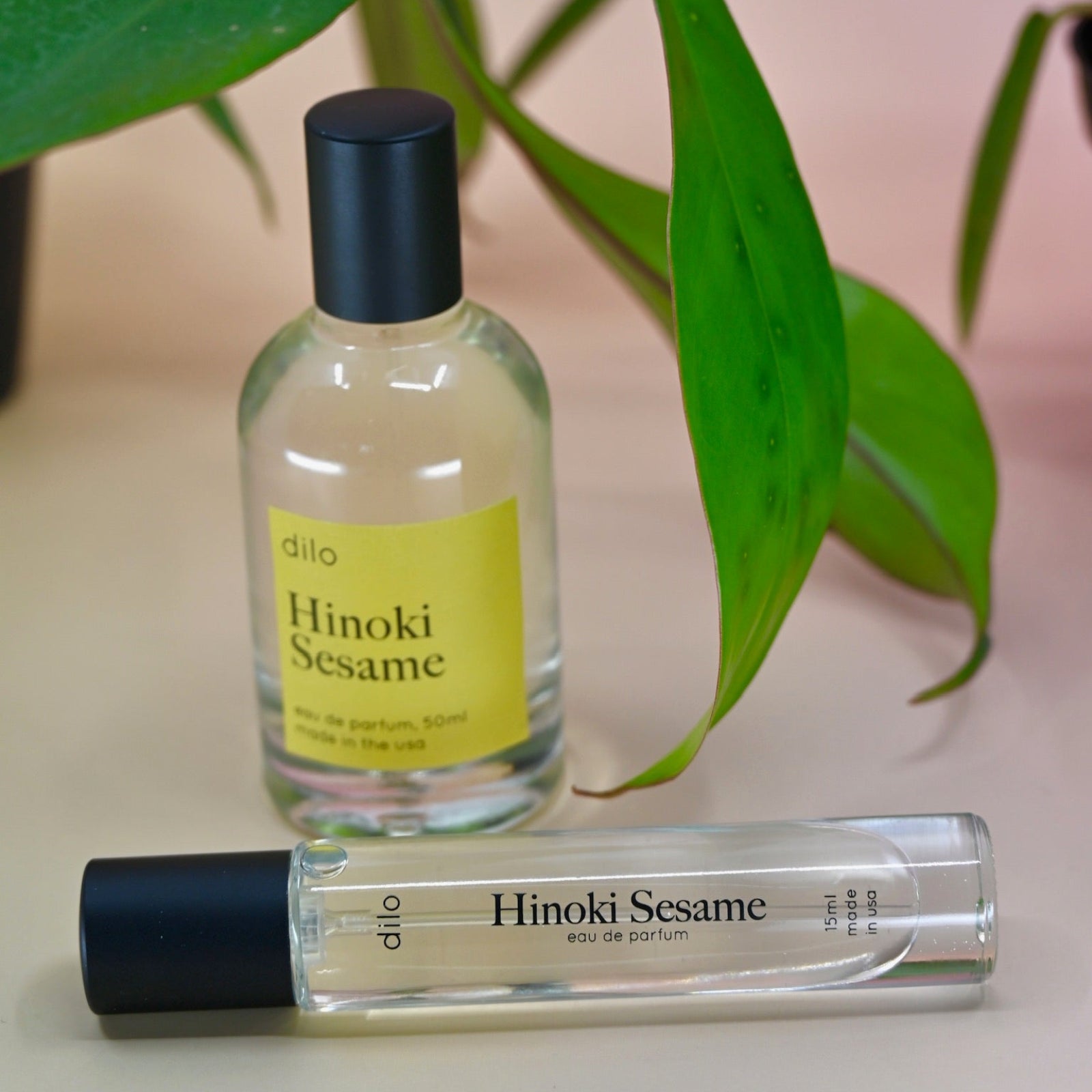The Hinoki Sesame 5ml Unisex Eau de Parfum regular sized bottle and the Travel Sprayer on a pink background with leaves.