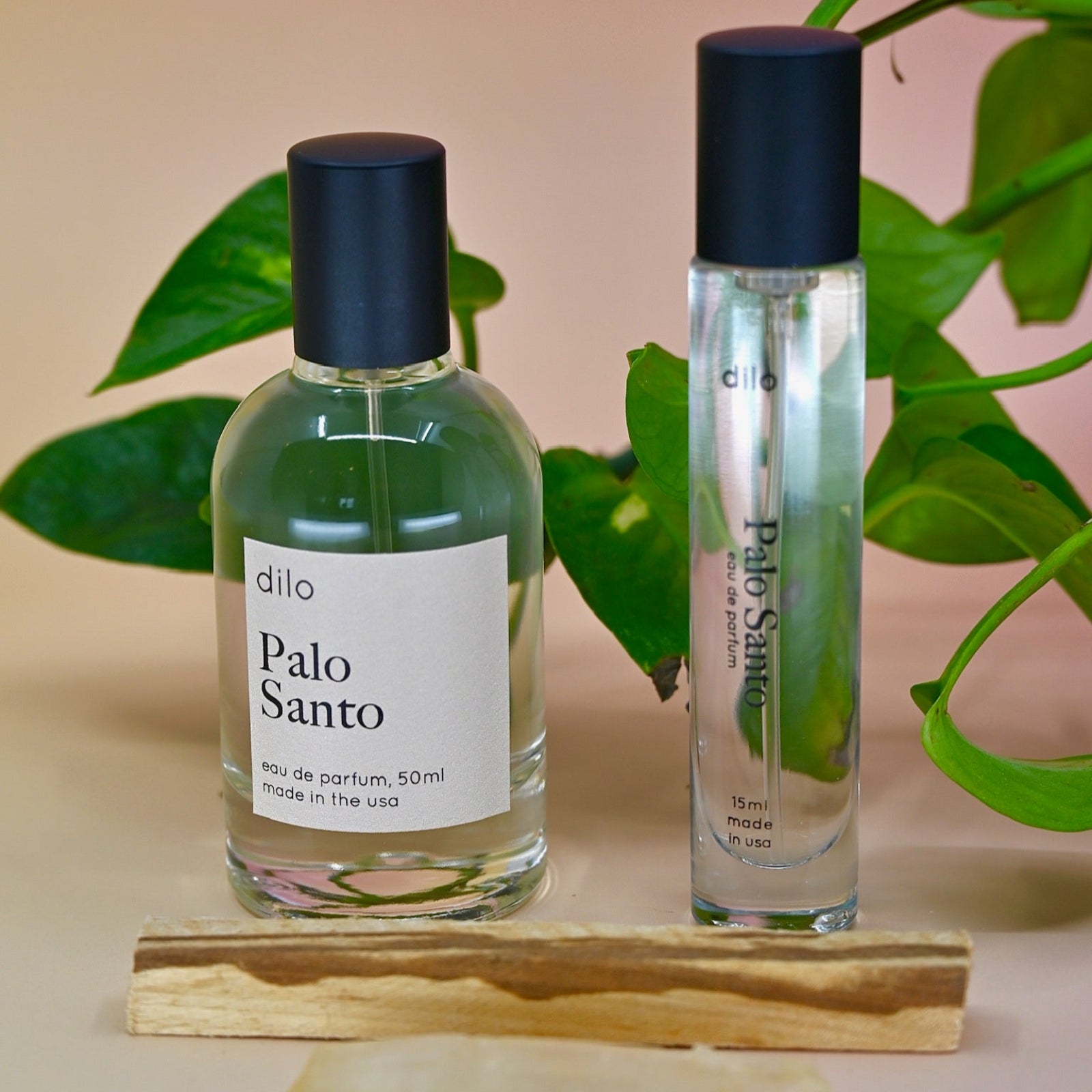 The Palo Santo 15ml Unisex Eau de Parfum Travel Sprayer and the regular size on a light pink background with leaves in the back and a stick of palo santo in front of the bottles.