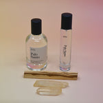 The Palo Santo 15ml Unisex Eau de Parfum Travel Sprayer and the regular size on a light pink background with a stick of palo santo in front of the bottles.