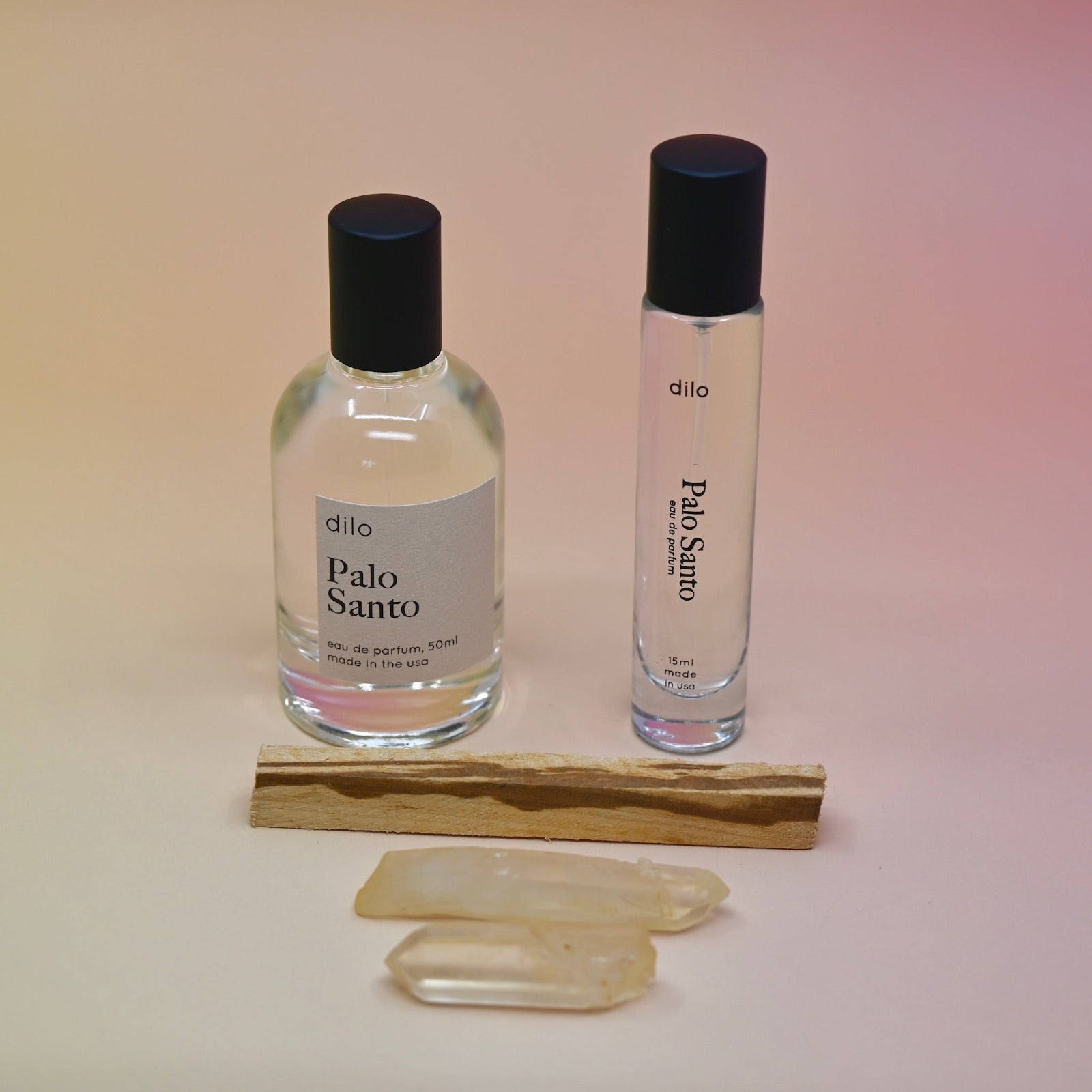 The Palo Santo 15ml Unisex Eau de Parfum Travel Sprayer and the regular size on a light pink background with a stick of palo santo in front of the bottles.