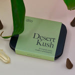 The Desert Kush Incense Cones - Pack of 25 on surrounded by crystals, incense cones, and leaves on a light pink background.