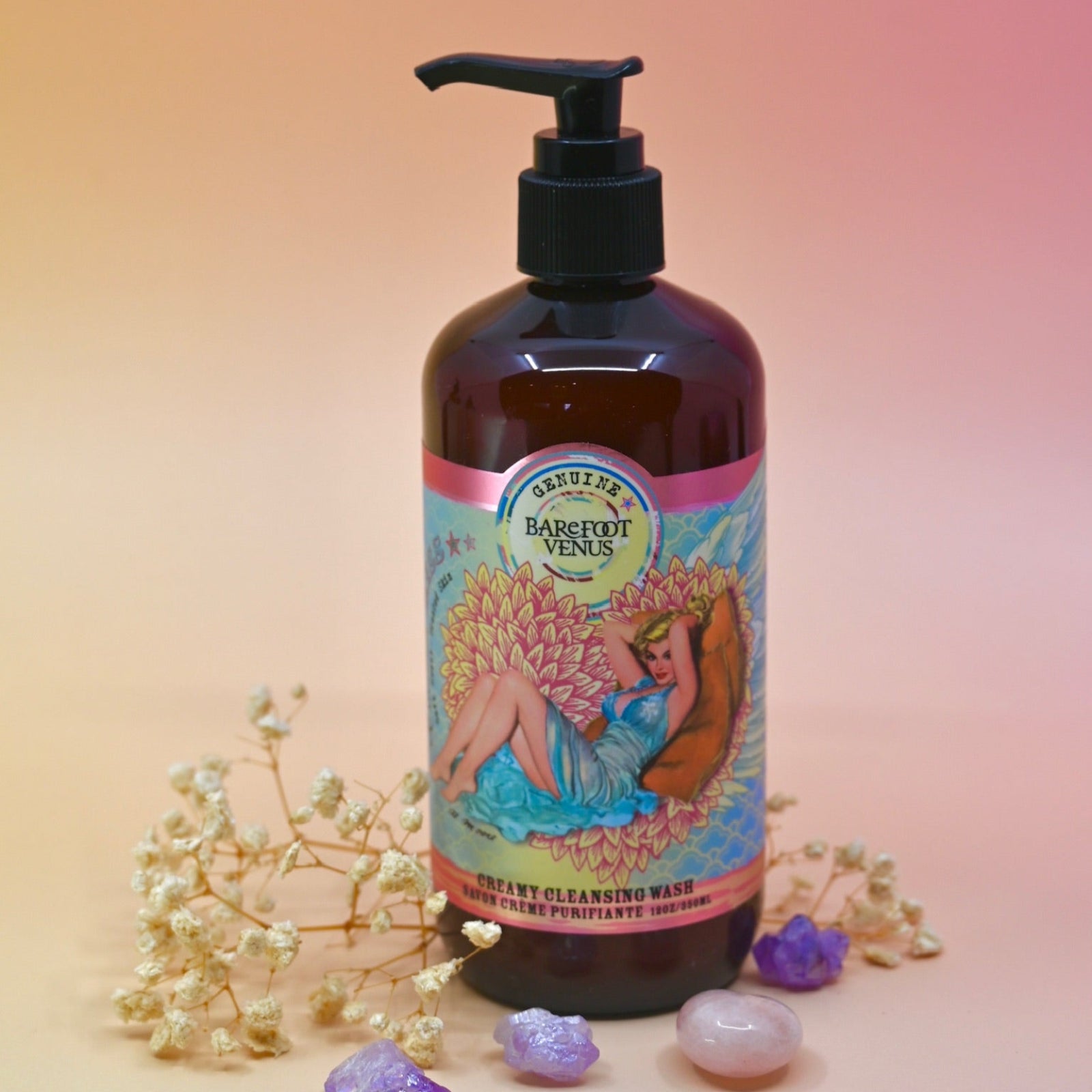 The Coconut Kiss Cleansing Hand & Body Wash surrounded by crystals and dried flowers on a light pink background.